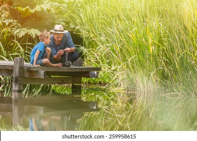 Young Father Teaching His Son To Make A Fishing Rod, They Are Sitting On A Wood Pontoon On A River