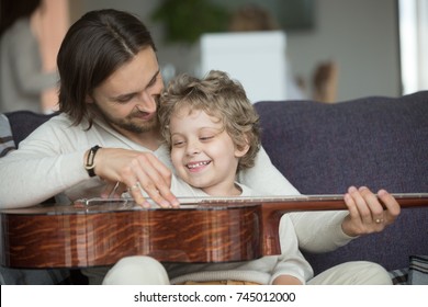 Young father teaching little son to play musical instrument at home, loving dad strumming guitar with small boy, parent and child relaxing on sofa, family leisure music activity, musically gifted kid