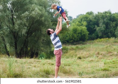 Young Father Throwing Up His Little Son Into Air And Catching Him Into Hands Outdoor. Dad Playing With His Child At Nature. Happy Family Spending Time Together Outside At Meadow. Beautiful Background