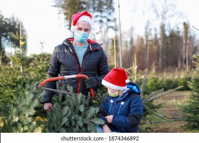 Young Father Wearing Surgical Face Mask With A Saw In His Hand And Little Son At Cut Your Own Christmas Tree Farm During Coronavirus, COVID-19 Lockdown. New Normal Life Holiday Celebration.