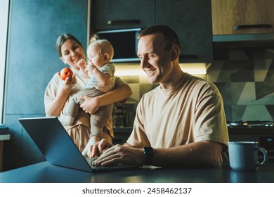 A young father works on a laptop while his wife takes care of the child. Work from home. A young couple in the kitchen with a child - Powered by Shutterstock