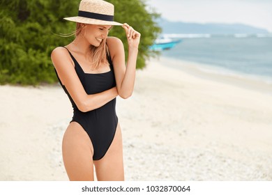Young female adventurer in black bathing suit, has slender legs and perfect body shape, wears straw hat, stands back to tropical ocean view with vegetation, comtemplates wonderful landscapes.