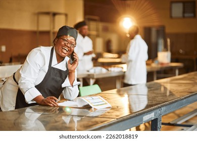 young female african chef wearing full chefs outfit and a black apron holding cellphone in industrial kitchen at night - Powered by Shutterstock