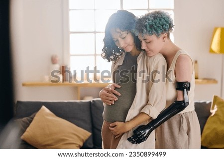 Young female amputee embracing her pregnant girlfriend and touching her belly while both standing in front of camera against couch