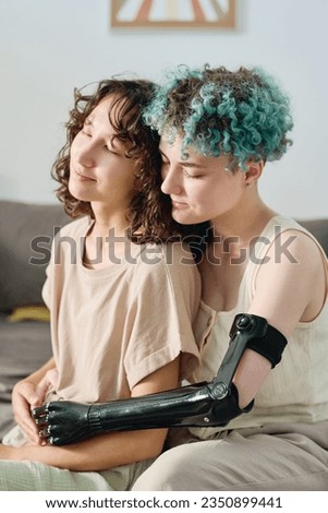 Young female amputee with short blue curly hair embracing her pretty brunette girlfriend while sitting close to her in front of camera