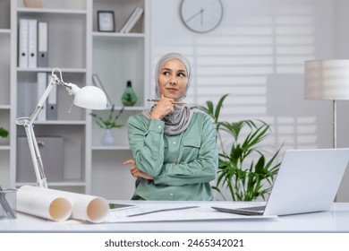 Young female architect thinking inside office, woman in hijab working with blueprints and layouts, using laptop at work. - Powered by Shutterstock