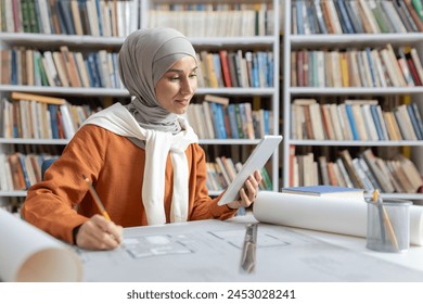 A young female architect wears a hijab while focusing on her project designs using a digital tablet in a library setting, surrounded by books.