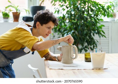 Young female artisan concentrated on modeling jug from raw clay while pottery lesson or workshop in creative studio. Woman ceramic business owner making craft for sale in handmade potter retail store