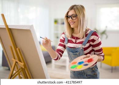 Young female artist painting on a canvas with a paintbrush and holding a color palette at home - Powered by Shutterstock