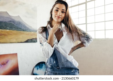 Young female artist standing in front of her paintings. Confident female painter looking at the camera in her art studio. Creative young woman displaying a collection of her artwork.