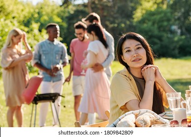 Junge weibliche asiatische Volksgruppe sitzt am Tisch vor der Kamera auf dem Hintergrund ihrer interkulturellen Freunde, die Grill machen