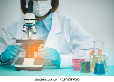 Young Female Asian Scientist With Mask Is Looking At The Glass Slide On Microscope And Clinician Studying Chemical Element In Medical Laboratory. Testing For Coronavirus Covid19 Vaccine