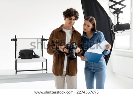 Young female assistant of professional photographer with documents discussing details of video or photo shooting, standing in photostudio and preparing for photoshoot