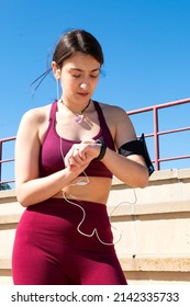 A Young Female Athlete Dressed In A Purple Top And Maroon Leggings Wearing A Cell Phone Bracelet, A Heart Pendant And Headphones Is Manipulating Her Smart Tracker In The Stands Of A Sports Area.