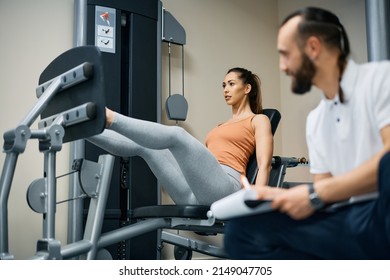 Young Female Athlete Exercising On Leg Press Machine While Having Sports Training With Fitness Instructor In Health Club.