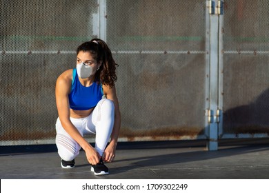 Young Female Athlete With Face Mask For Protecting Against Covid-19 Contagion Getting Ready For Urban Running And Fitness Workout. Motivated Woman Training Outside Under Coronavirus Health Crisis.