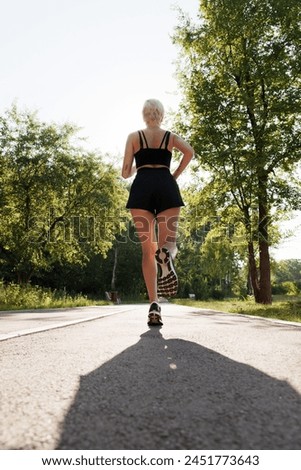 A young female athlete is running on a sunlit path surrounded by trees, showcasing a healthy lifestyle and fitness.