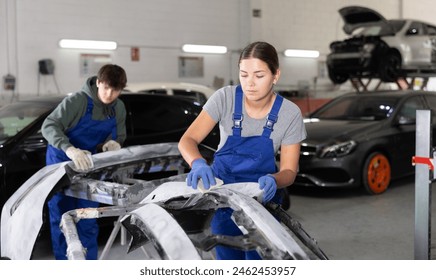 Young female auto mechanic in blue overalls preparing car for painting, sanding removed bumper with sandpaper after applying putty in specialized garage, focused on work - Powered by Shutterstock