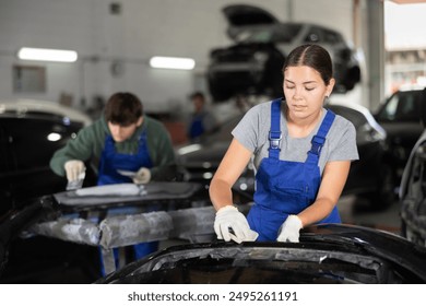 Young female auto mechanic in blue overalls preparing car for painting, sanding removed bumper with sandpaper after applying putty in specialized garage, focused on work - Powered by Shutterstock