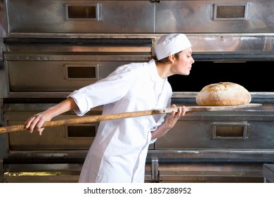 Young Female Baker Taking Out A Loaf Of Bread From The Oven And Smelling It While Holding It In A Wooden Tray