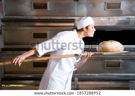 Young female baker taking out a loaf of bread from the oven and smelling it while holding it in a wooden tray