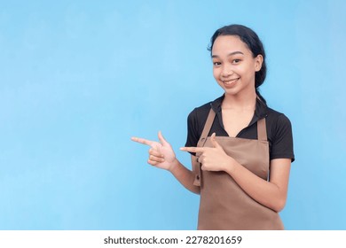 A young female barista of asian descent wearing a brown apron and black shirt finger pointing something to the left. Against a light blue background. - Shutterstock ID 2278201659