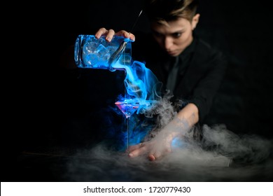 young female barman professionally pours illuminated smoky cocktail from glassy mixing bowl with steel strainer into martini glass with ice. A lot of smoke in bar.