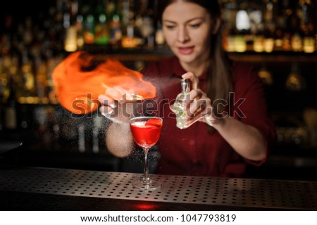 Young female bartender sprinkling a cocktail glass with tasty Aperol syringe cocktail with a peated whisky and making a smoky note
