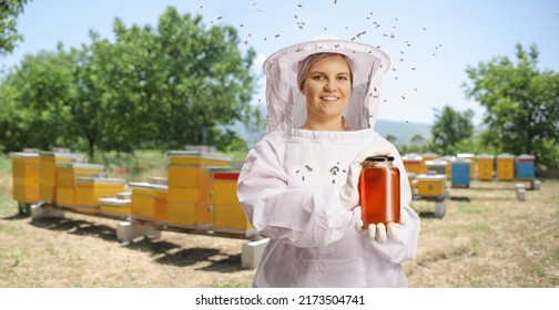 Young female bee keeper in a uniform holding a jar of honey on apiary and bees flying