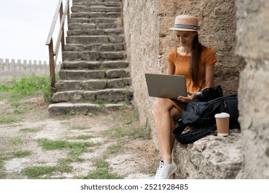 Young female blogger sits by ancient stone wall editing photos on her laptop. She wears a summer dress and hat, documenting her travels through medieval fortresses and historical landmarks. - Powered by Shutterstock