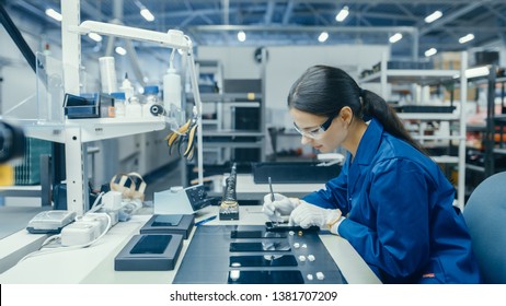 Young Female In Blue Work Coat Is Assembling Printed Circuit Boards For Smartphones. Electronics Factory Workers In A High Tech Factory Facility.