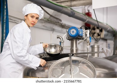 Young Female Brewmaster In White Uniform Working In Small Brewery, Pouring Hop Pellets From Bowl Into Beer Production Fermenter