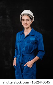 Young female builder in hardhat and blue coveralls keeping hands in pockets while standing in front of camera in isolation