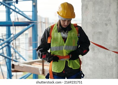 Young Female Builder In Protective Helmet And Reflective Vest Fastening Safety Belt On Waist Before Building Work At Construction Site