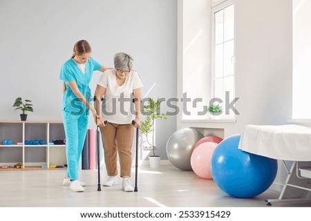 Young female caregiver, nurse helping elderly woman to walk with crutches during rehabilitation in clinic. Physiotherapist providing assistance and supporting senior patient after injury or stroke.