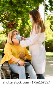 Young Female Caregiver Wearing A Surgical Mask Adjusting The Surgical Mask Of A Mature Woman In A Wheelchair On An Out Of Focus Background. Disability And Safety Concept.