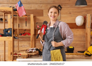 Young female carpenter with chain, bolt cutter and USA flag in workshop. Labor Day celebration - Powered by Shutterstock