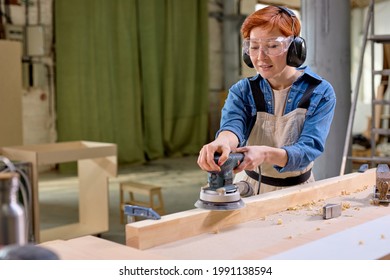 Young Female Carpenter Working With Wood Using Electric Sander In Workshop. Joinery Work On The Production And Renovation Of Wooden Furniture. Small Business Concept