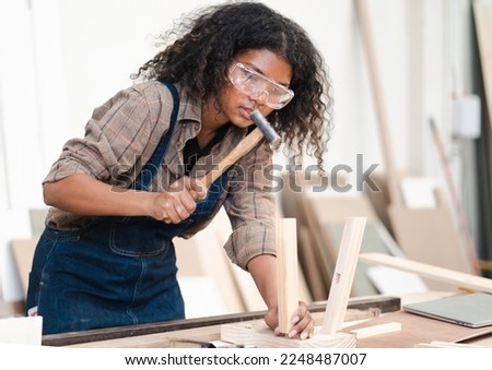 Young female carpenter is working in woodwork furniture in carpentry workshop. Multiracial woman woodworker busy using hammer to assemble DIY woodworking at workbench. Joiner and craftsman person.