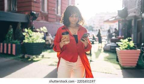 Young Female In Casual Clothes With Cup Of Hot Drink Standing On Street And Browsing Mobile Phone While Looking At Screen In Back Lit