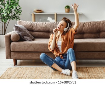 Young female in casual outfit listening to music in headphones and singing while sitting on floor near sofa and having fun at home