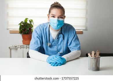 Young female caucasian General Practitioner sitting by her desk in office,wearing blue uniform,protective gloves  face mask,virtual tele visit via video call,GP doctor on demand telemedicine concept - Powered by Shutterstock