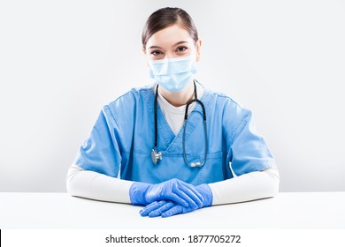 Young Female Caucasian General Practitioner Sitting At Office Desk,wearing Blue Uniform,protective Gloves,face Mask,virtual Tele Visit Via Video Call,UK GP Doctor On Demand, Telemedicine Concept