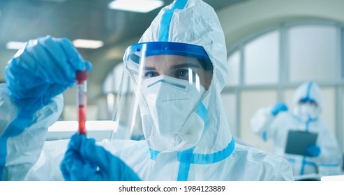 Young female Caucasian scientist in protective coverall, mask and face shield holding test tube with blood sample making research about covid virus in clinical laboratory and working on vaccine