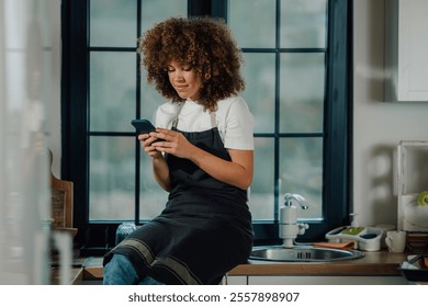 Young female chef with curly hair wearing apron sitting on kitchen counter using smartphone, taking break from cooking, checking online orders or social media in restaurant kitchen - Powered by Shutterstock