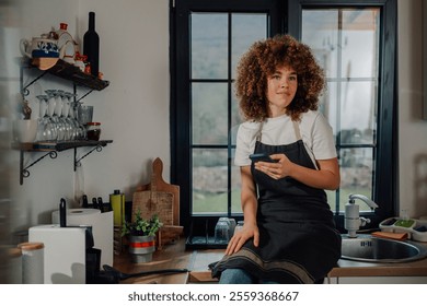 Young female chef with curly hair wearing apron sitting on kitchen counter using smartphone and looking away after finishing work in restaurant kitchen - Powered by Shutterstock