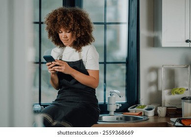 Young female chef with curly hair is sitting on the kitchen counter, using a smartphone and wearing an apron in a modern restaurant kitchen - Powered by Shutterstock