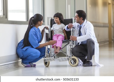 Young Female Child Patient In Wheelchair Sitting In Hospital Corridor With Indian Asian Female Nurse And Male Doctor