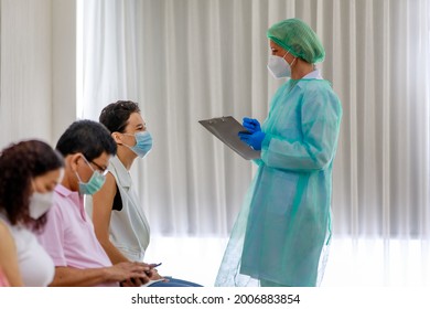 Young female citizen wears face mask in Coronavirus vaccinating queue line sitting checking her personal information from paper board in hand of full protective suit nurse who standing nearby.