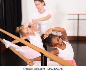 Young female classical dance teacher helping her little students near ballet barre in dancing hall